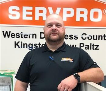 A male employee standing in front of the SERVPRO Orange House logo, wearing a SERVPRO shirt
