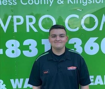 A smiling male employee standing in front of a SERVPRO green trucks wearing a black SERVPRO polo shirt