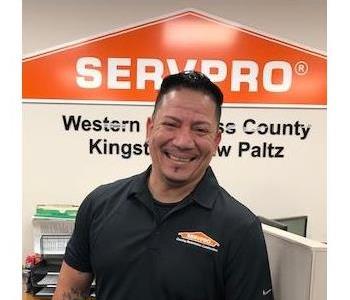 A smiling male employee in a black shirt standing in front of the SERVPRO Orange House Logo