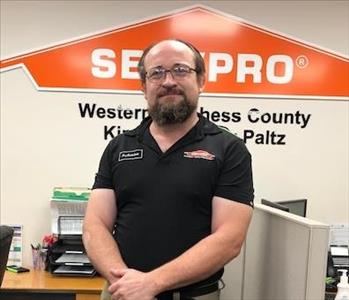 A smiling male employee in a black shirt standing in front of the SERVPRO Orange House Logo