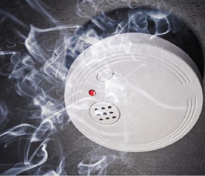 An image of a white smoke detector on a black wall surrounded by wisps of smoke