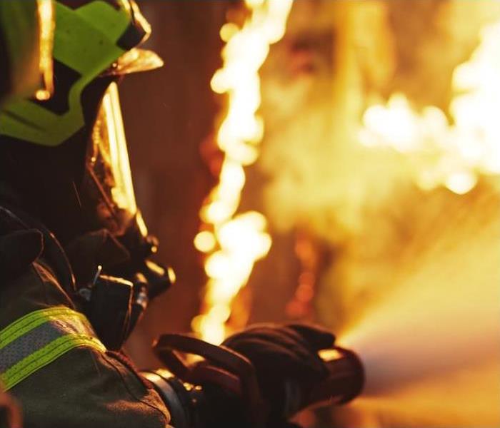 A firefighter holding a fire hose shooting water at a wall of flames.