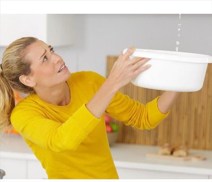 A woman in a yellow shirt holding a white bucket above her head to catch water dripping from the ceiling.