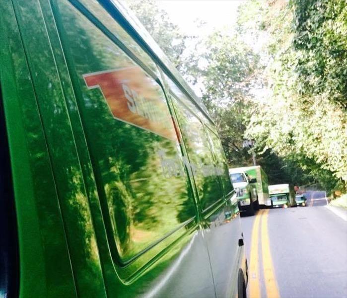A fleet of green SERVPRO vehicles behind each other, viewed from the side mirror of the lead truck