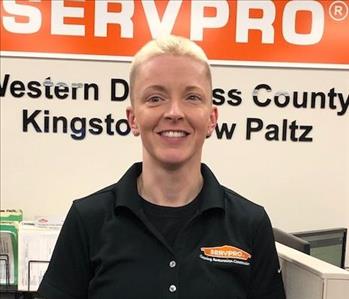 A smiling female employee wearing a SERVPRO shirt and standing in front of the SERVPRO Orange House logo